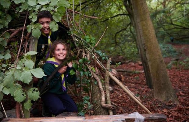 Two cub scouts one male and one female are inside a woodland den and smiling the den is made out of trees logs and leaves and they look very proud